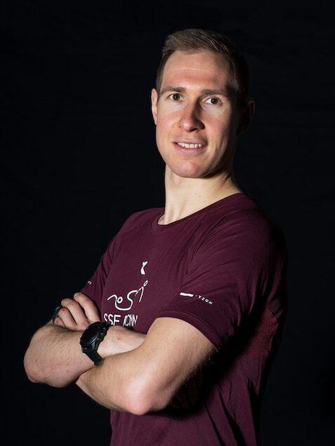 Portr&auml;t eines l&auml;chelnden Mannes in einem burgunderfarbenen T-Shirt, mit verschr&auml;nkten Armen vor schwarzem Hintergrund.