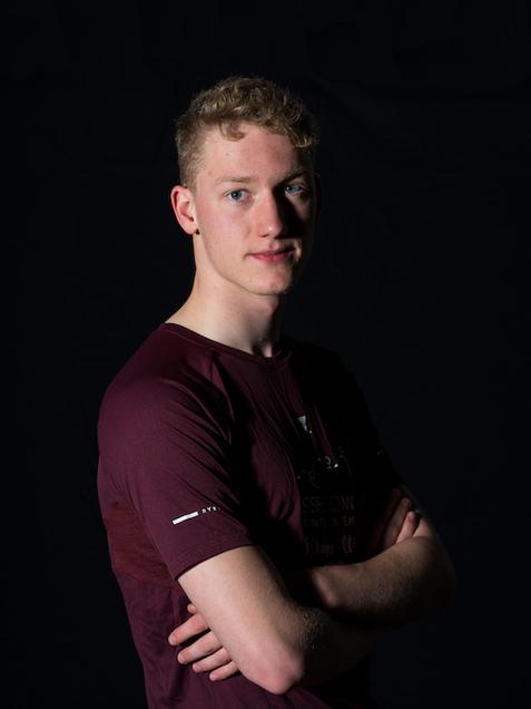 Junger Mann mit Kurzhaarschnitt, in einem burgunderfarbenen T-Shirt, steht mit verschr&auml;nkten Armen vor dunklem Hintergrund.