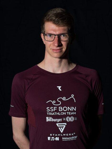 Portr&auml;t eines Mannes in einem roten Triathlon-Shirt mit Logo des SSF Bonn Triathlon Teams, vor schwarzem Hintergrund.