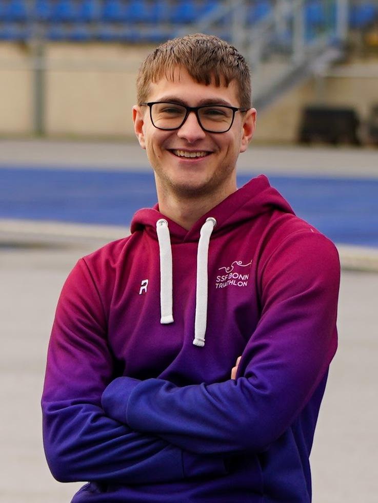 L&auml;chelnder junger Mann in lila-rot gradientem Hoodie, steht mit verschr&auml;nkten Armen auf einem Sportgel&auml;nde.