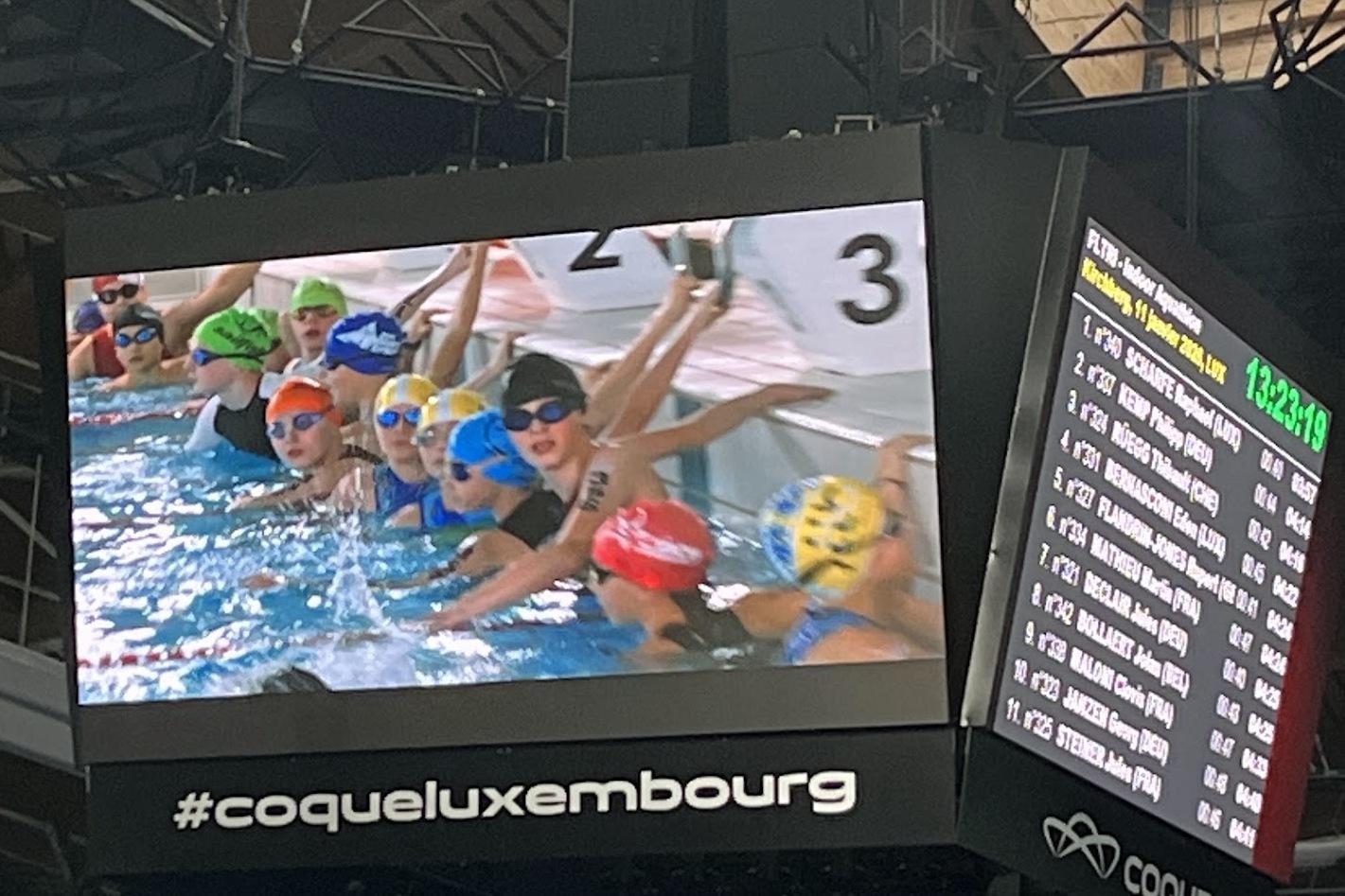 Schwimmerinnen in Badekappen und Schwimmbrillen starten in einem Wettkampf vor einer digitalen Anzeigetafel.