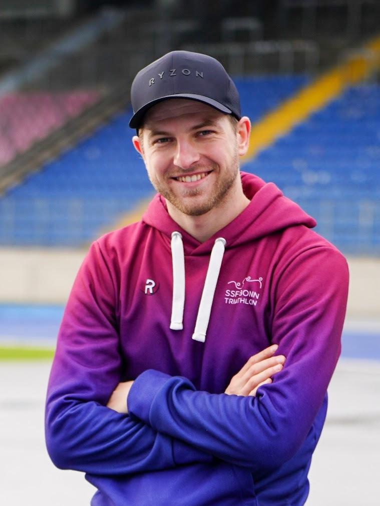 Junger Mann mit kurzgeschnittenem Haar l&auml;chelt in lila-rot gestreifter Kapuzenjacke vor einem Sportstadion.