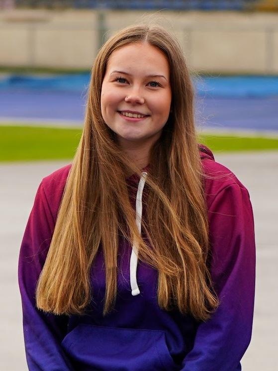 Junge Frau mit langen Haaren sitzt an einem Sportplatz, tr&auml;gt einen lila-rot gef&auml;rbten Hoodie und l&auml;chelt.