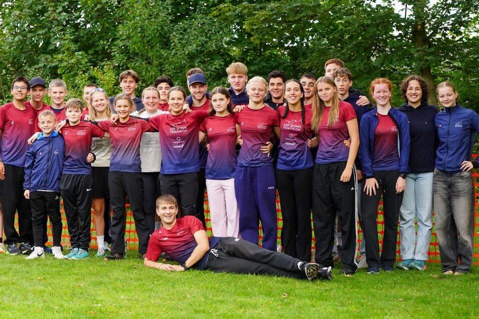 Gruppe von Jugendlichen in einheitlichen Sportshirts, l&auml;chelnd, auf einer Wiese vor gr&uuml;ner Kulisse.