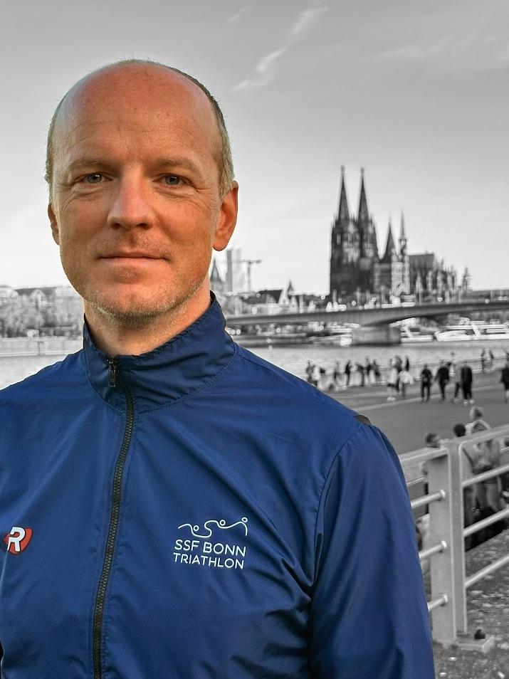 Portr&auml;t eines Mannes in blauer Triathlonjacke vor unscharfem Hintergrund mit K&ouml;lner Dom und Menschen am Rheinufer.