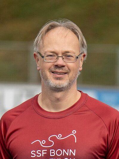 Mann mit Brille und leichtem Bart l&auml;chelt in einem roten Shirt mit SSF Bonn-Logo vor einem Sportplatz.