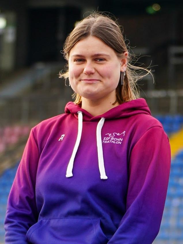 Junge Frau mit kurzen Haaren in lila-rot gef&auml;rbtem Hoodie steht in einem Stadion mit leeren Trib&uuml;nen im Hintergrund.