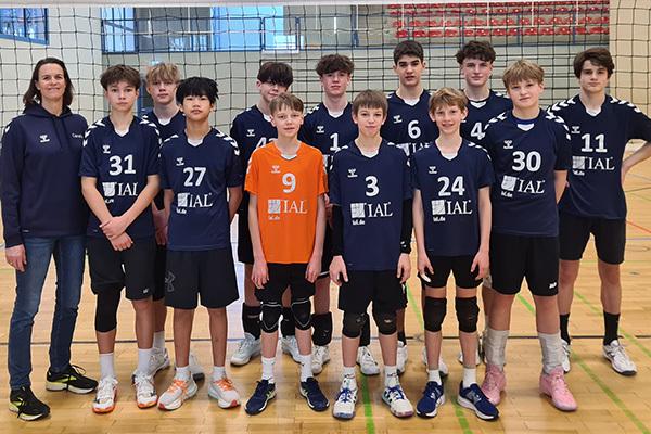 Gruppensfoto einer Volleyballmannschaft mit 12 Jungen und einer Trainerin, auf einem Volleyballplatz in der Halle.