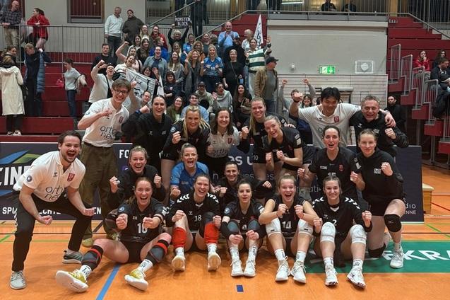 Feiernde Volleyballmannschaft posiert in triumphierender Haltung vor jubelndem Publikum in der Sporthalle.
