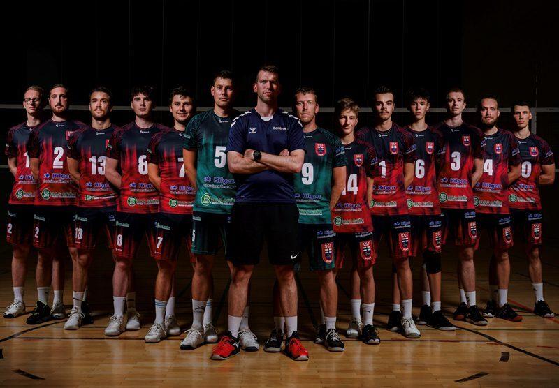 Aufstellung einer Volleyballmannschaft mit Trainer, alle in einheitlichen Trikots, auf einem Volleyballfeld.