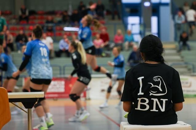 Zuschauerin mit T-Shirt &bdquo;I &hearts; BNN&ldquo; sitzt am Rande eines Volleyballspiels mit aktiven Spielerinnen im Hintergrund.