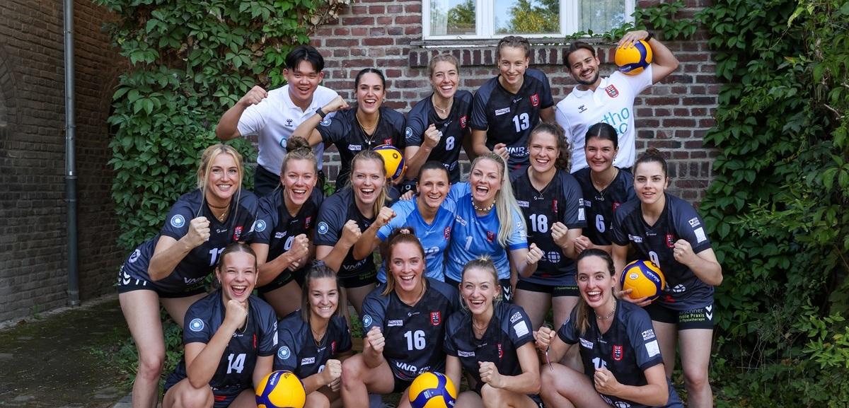 Gruppenfoto eines Volleyballteams mit 15 Spielerinnen, die in sportlicher Kleidung mit B&auml;llen vor einer Hauswand posieren.