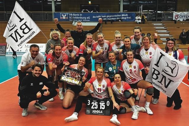 Feiernde Volleyballmannschaft mit Trainer, zwei Flaggen und einem Pokal auf dem Spielfeld nach dem Sieg.