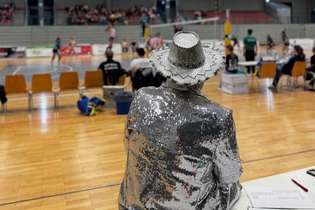 Person in glitzerndem Anzug beobachtet ein Volleyballspiel in einer Halle, im Hintergrund Spieler und Zuschauer.