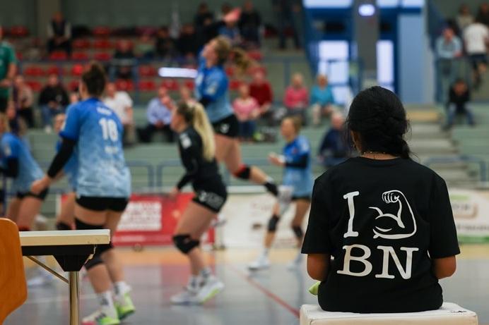 Zuschauerin mit T-Shirt &bdquo;I &hearts; BNN&ldquo; sitzt am Rande eines Volleyballspiels mit aktiven Spielerinnen im Hintergrund.