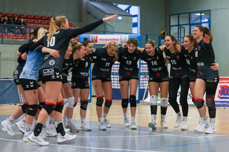 Zehn Sportlerinnen feiern im Kreis auf dem Volleyballfeld, in einheitlicher Teamkleidung, Stimmung ist ausgelassen.
