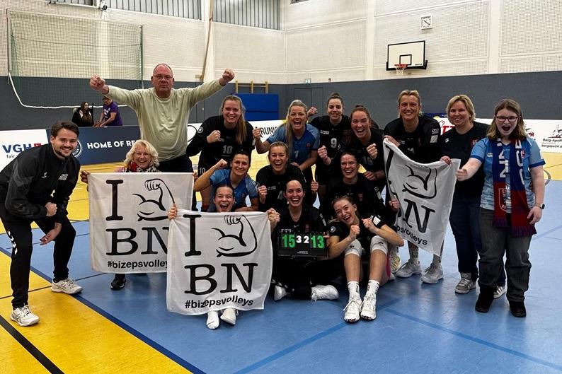 Feiernde Volleyballmannschaft mit Fans, gehaltenen Schildern und Punktzahl 15:32:13 in einer Sporthalle.