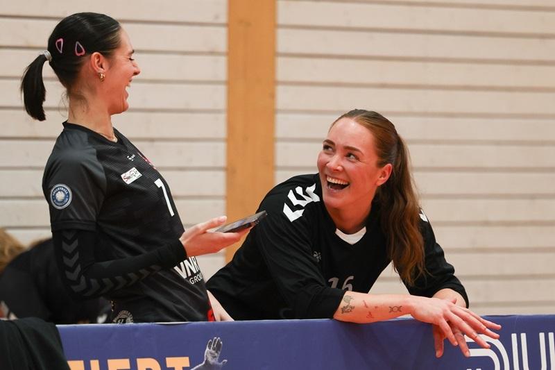 Zwei lachende Handballspielerinnen sitzen an der Bank, die eine h&auml;lt ein Smartphone in der Hand.