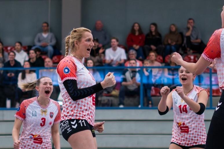 Vier Volleyballspielerinnen jubeln lautstark auf dem Spielfeld, im Hintergrund ist ein Publikum zu sehen.