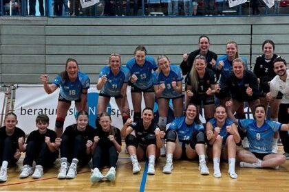 Gruppierung von Handballspielerinnen in Trikots, posierend mit F&auml;usten, vor jubelndem Publikum mit Fahnen.