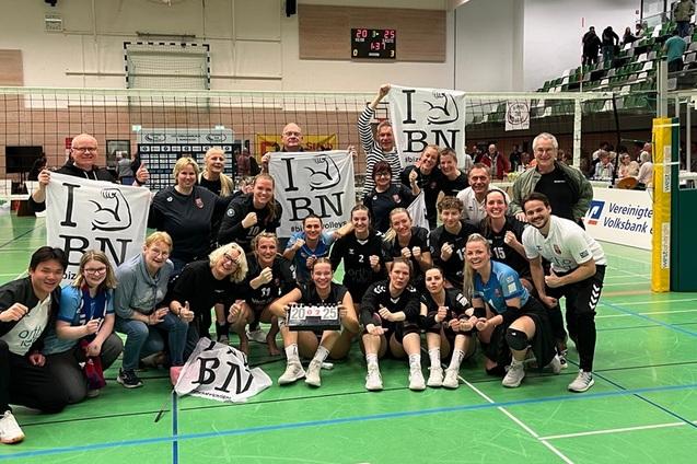 Teamfoto der Volleyballmannschaft mit Fans, die Fahnen halten, vor einem Netz in einer Sporthalle nach einem Spiel.