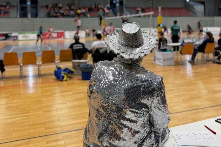 Person in glitzerndem Anzug beobachtet ein Volleyballspiel in einer Halle, im Hintergrund Spieler und Zuschauer.