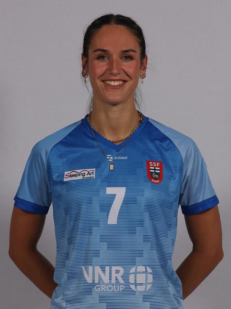 L&auml;chelnde Sportlerin in hellblauem Trikot mit der Nummer 7, vor neutralem Hintergrund, mit kurzen Haaren und Schmuck.