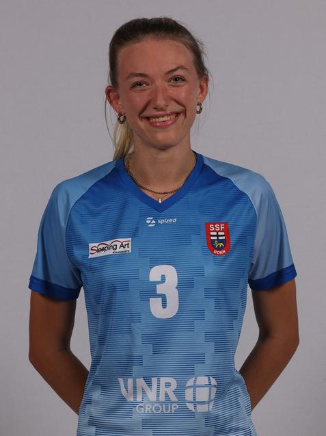 Portr&auml;t einer sportlichen Frau in einem blauen Trikot mit der Nummer 3 und Logos auf der Brust, l&auml;chelnd vor neutralem Hintergrund.