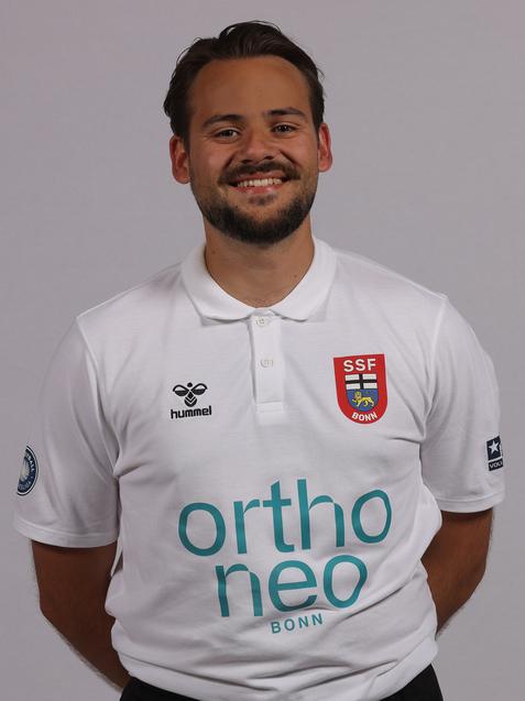 L&auml;chelnder Mann in wei&szlig;em Polohemd mit Vereinslogo und Sponsorenaufdruck, vor neutralem Hintergrund.