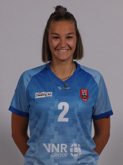 Portrait einer sportlichen Frau in blauem Trikot mit der Nummer 2, l&auml;chelnd vor neutralem Hintergrund.