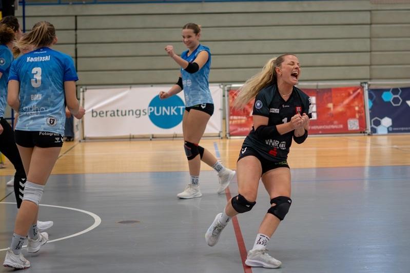 Drei Volleyballspielerinnen jubeln auf dem Spielfeld, eine Spielerin schreit vor Freude, im Hintergrund Werbung.