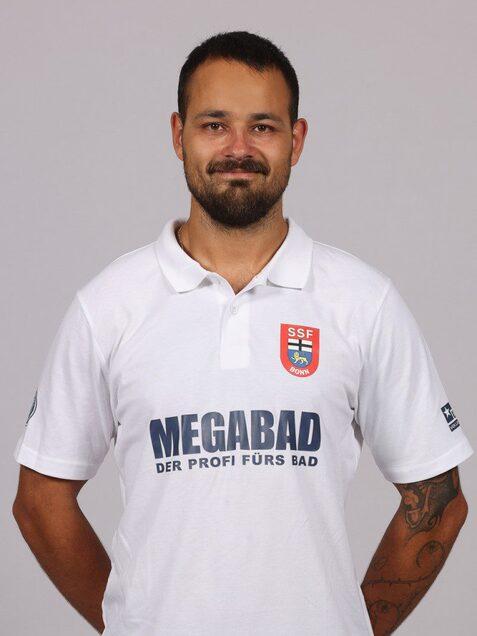 Portrait eines Mannes in einem wei&szlig;en Poloshirt mit Logos, l&auml;chelnd vor einem neutralen grauen Hintergrund.