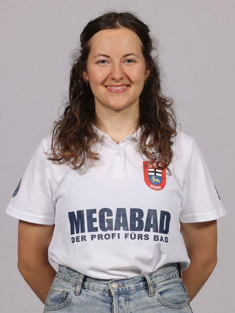 Portr&auml;t einer l&auml;chelnden Frau mit langen, lockigen Haaren in einem wei&szlig;en Poloshirt mit Aufdruck, vor einem grauen Hintergrund.