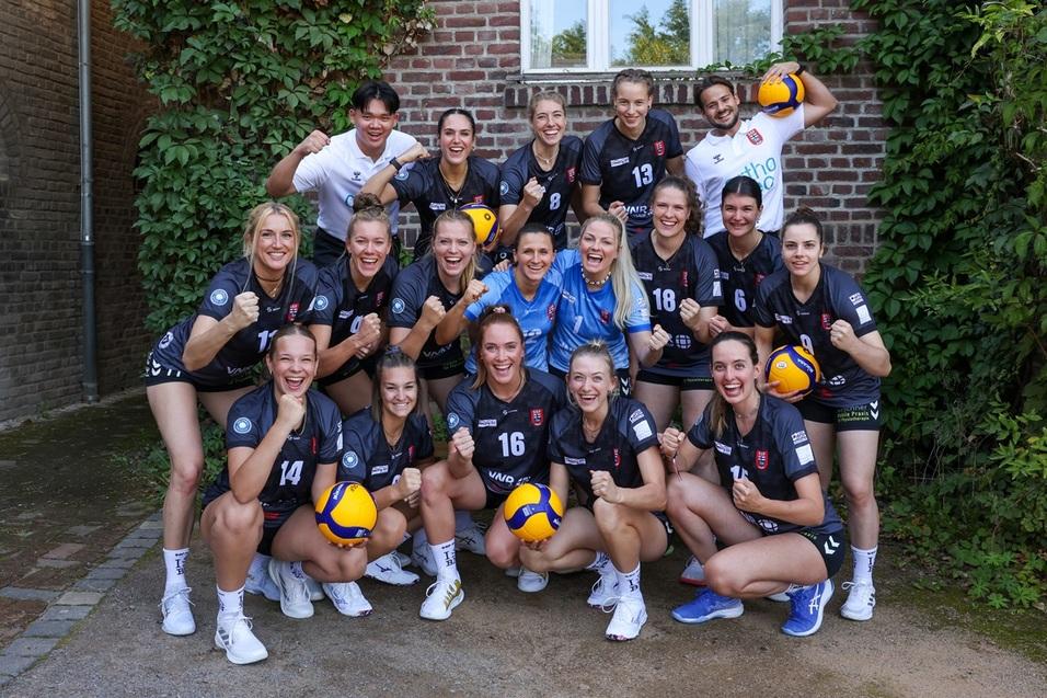 Gruppenfoto eines Volleyballteams mit 15 Spielerinnen, die in sportlicher Kleidung mit B&auml;llen vor einer Hauswand posieren.
