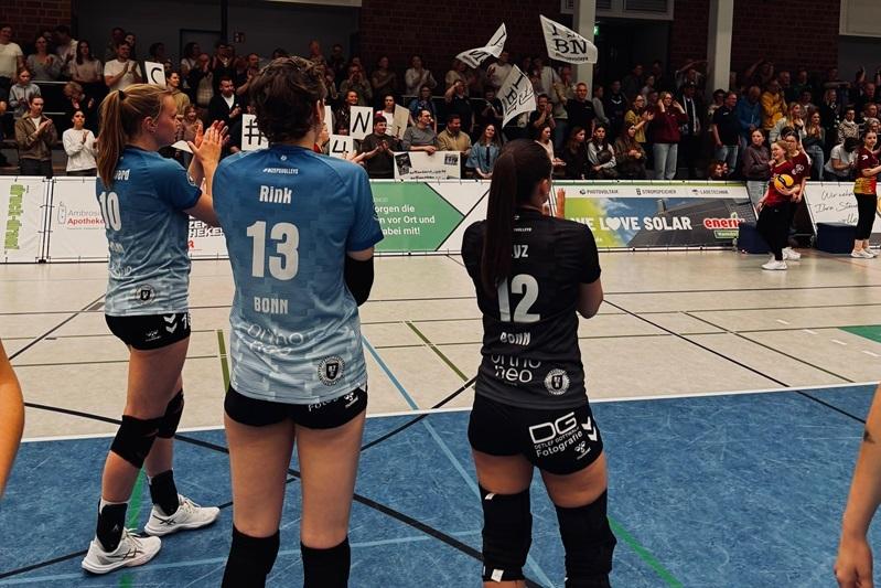 Drei Volleyballerinnen in Trikots stehen an der Spielfeldlinie, w&auml;hrend eine jubelnde Menge im Hintergrund das Spiel unterst&uuml;tzt.