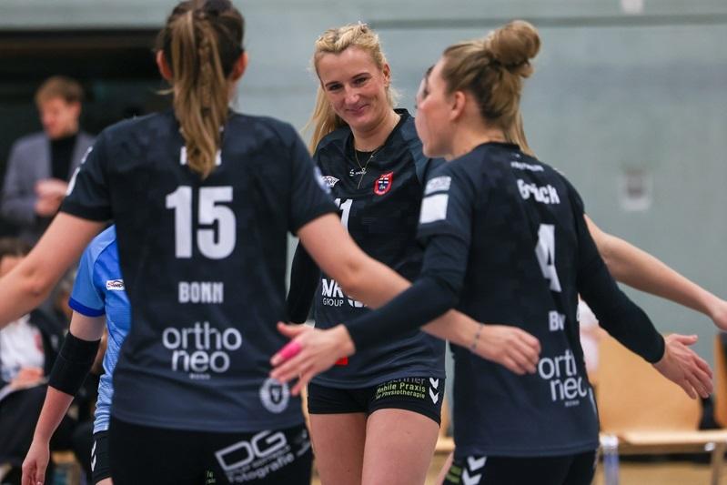 Drei weibliche Volleyballspielerinnen in schwarzen Trikots jubeln miteinander in einer Sporthalle.