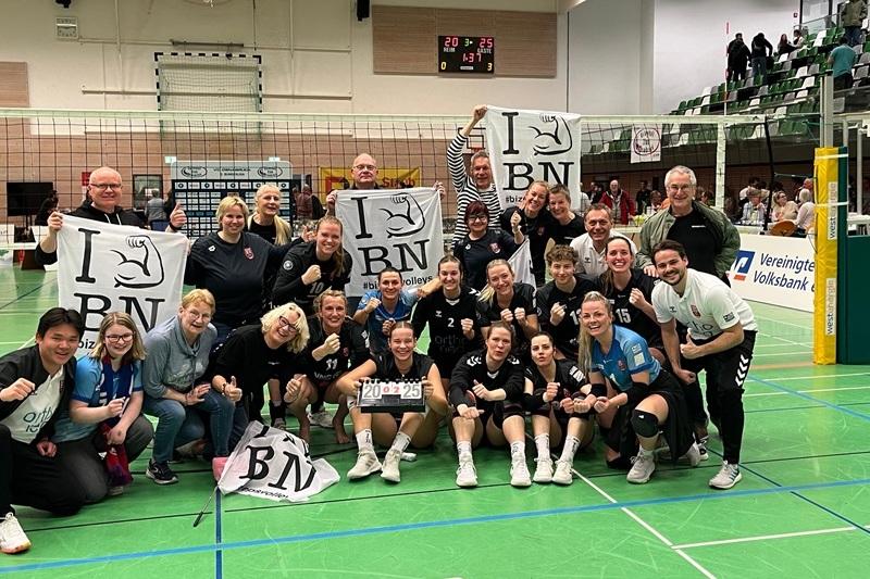 Teamfoto der Volleyballmannschaft mit Fans, die Fahnen halten, vor einem Netz in einer Sporthalle nach einem Spiel.