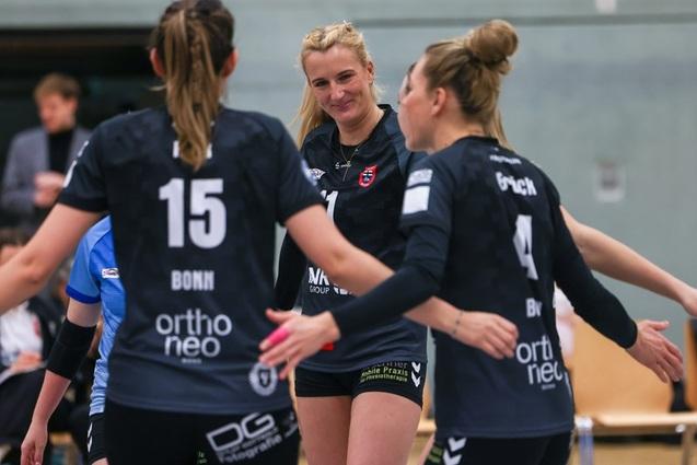 Drei weibliche Volleyballspielerinnen in schwarzen Trikots jubeln miteinander in einer Sporthalle.