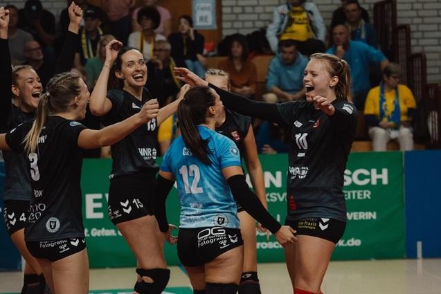Sechs Volleyballspielerinnen jubeln zusammen auf dem Spielfeld, w&auml;hrend Zuschauer im Hintergrund anfeuern.