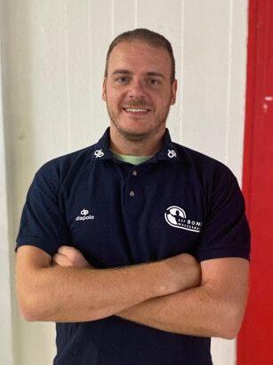 L&auml;chelnder Mann in Sportbekleidung mit verschr&auml;nkten Armen vor einer farbigen Wand.