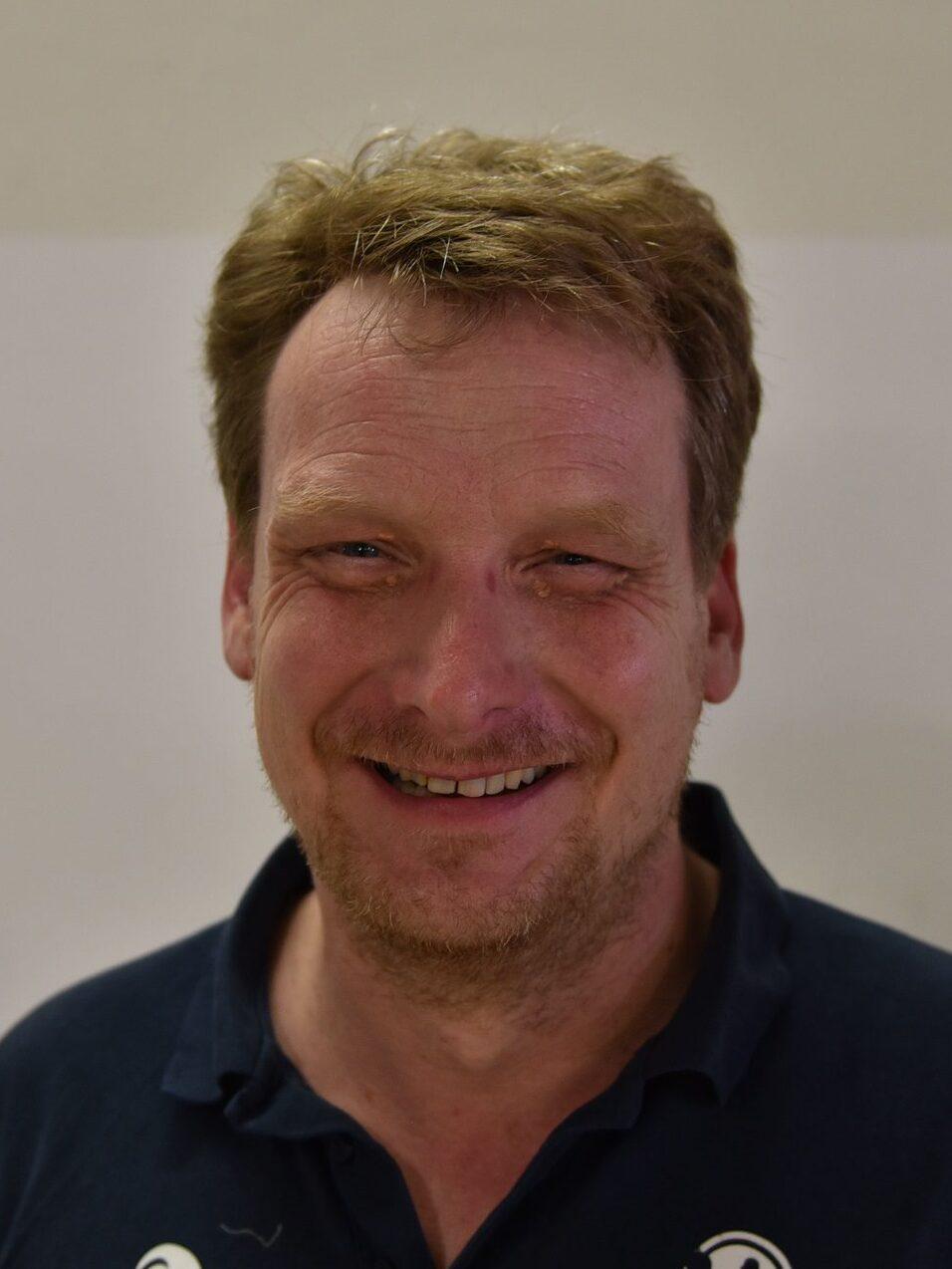 Portr&auml;t eines l&auml;chelnden Mannes mit kurzen, blonden Haaren, in einem dunklen Polohemd vor einer hellen Wand.