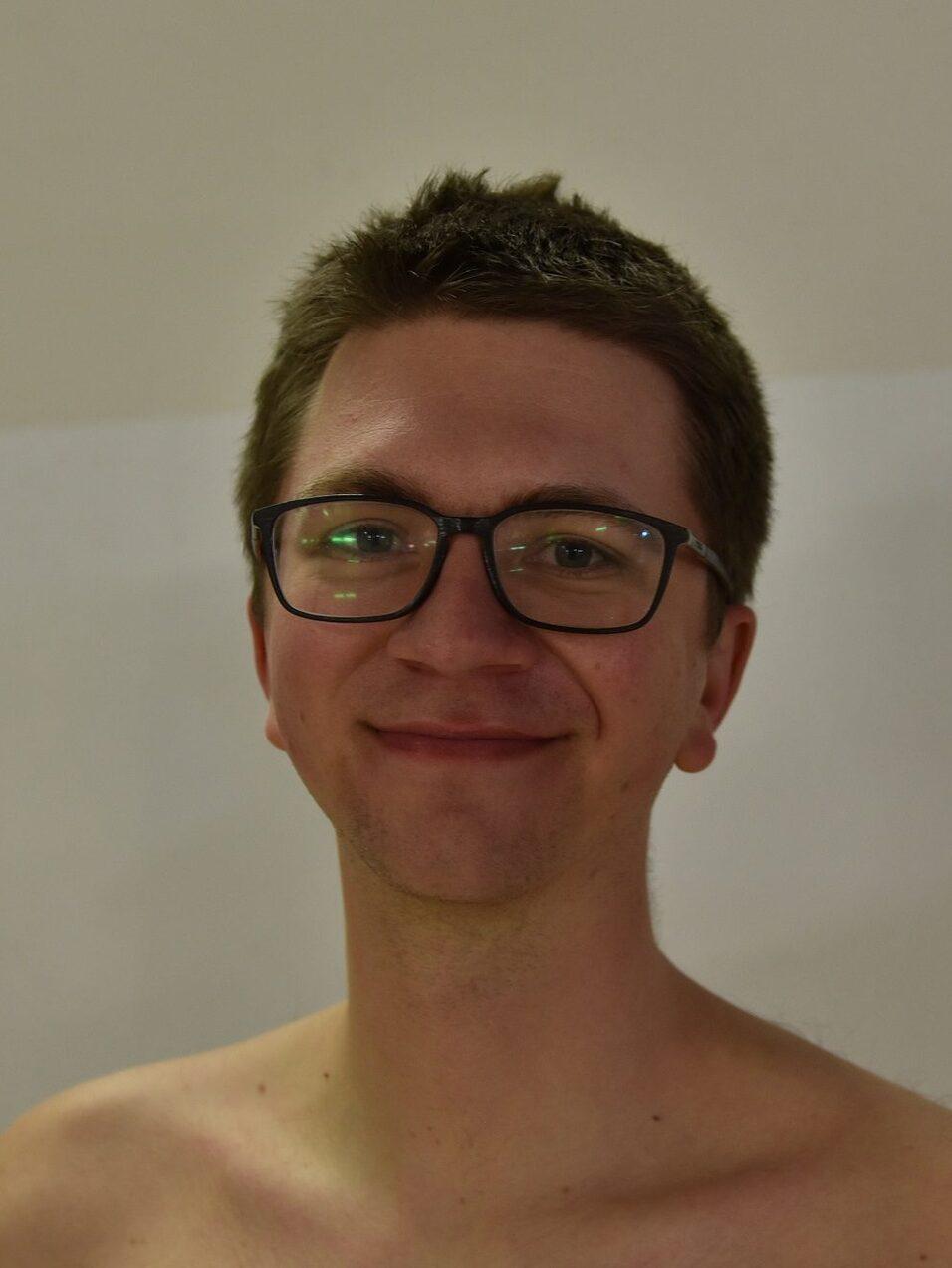 Junger Mann mit Brille, l&auml;chelnd vor einfarbiger Wand, ohne Oberbekleidung.