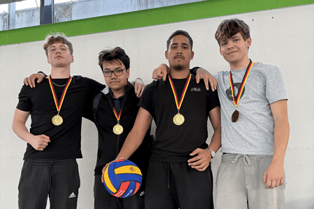Vier Jugendliche in sportlichen Outfits stehen gemeinsam, jeder tr&auml;gt eine Medaille und h&auml;lt einen Volleyball.