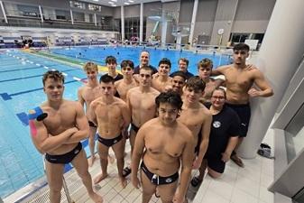 Gruppe von Schwimmern in Badebekleidung posiert am Beckenrand eines Schwimmbades mit blauem Wasser im Hintergrund.