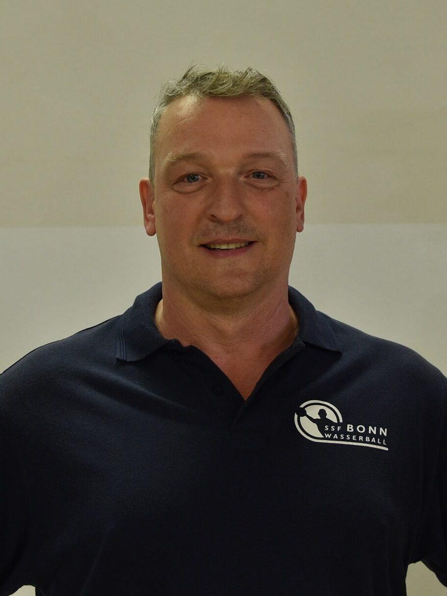 Mann in blauem Polo-Shirt mit Logo steht vor einer hellen Wand und l&auml;chelt in die Kamera.