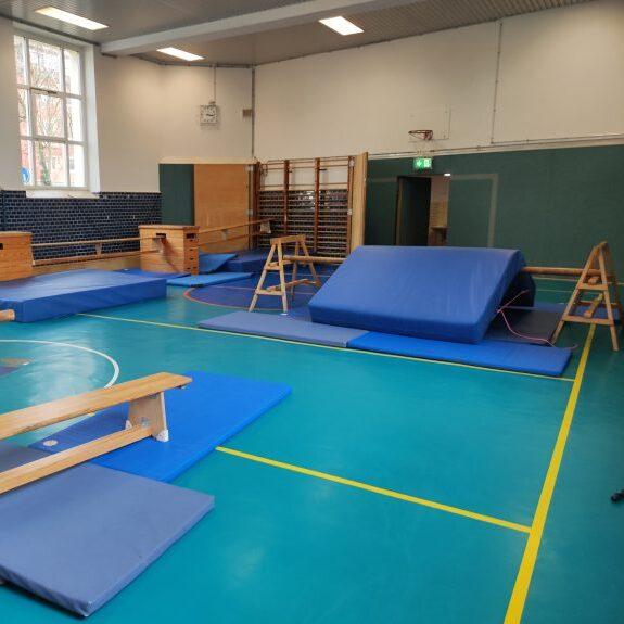 Sporthalle mit Turnger&auml;ten, blauen Matten und Barren auf einem gr&uuml;nen Boden, hell erleuchtet von gro&szlig;en Fenstern.