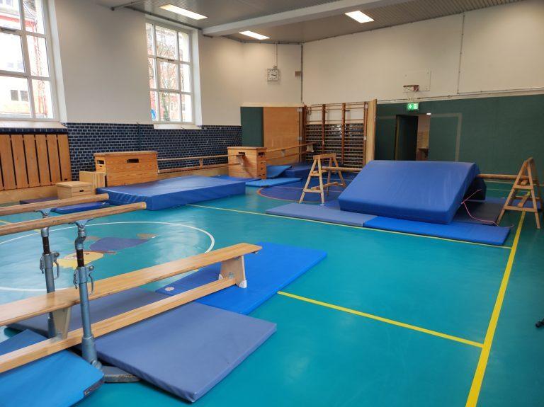 Sporthalle mit Turngeräten, blauen Matten und Barren auf einem grünen Boden, hell erleuchtet von großen Fenstern.