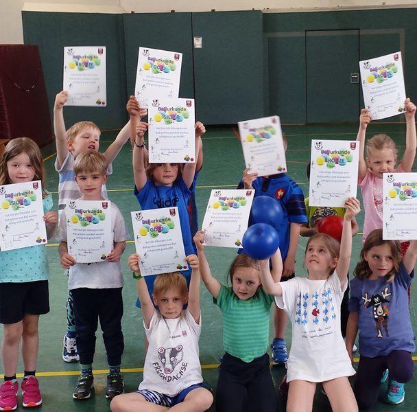 Gruppe von Kindern h&auml;lt Urkunden hoch, w&auml;hrend sie auf einem Sportboden stehen, einige mit B&auml;llen in den H&auml;nden.
