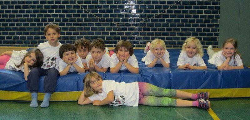 Gruppe von Kindern in wei&szlig;en T-Shirts, die auf einer blauen Matratze in einer Sporthalle liegen und posieren.