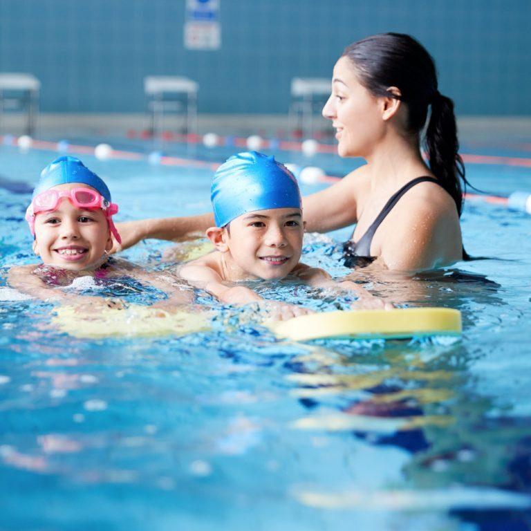 Zwei Kinder schwimmen im Wasser, w&auml;hrend eine Schwimmtrainerin sie mit einem Schwimmbrett unterst&uuml;tzt.